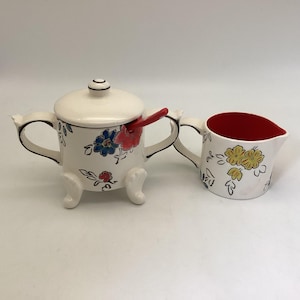 May include: A white ceramic sugar bowl with a lid and a matching creamer. The sugar bowl has a red spoon and is decorated with blue and red floral designs. The creamer has a red interior and yellow floral designs.