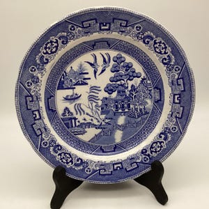May include: A blue and white ceramic plate with a Willow pattern design. The plate features a detailed landscape scene with pagodas, trees, a bridge, and birds. The rim has a decorative border. The plate is displayed on a black stand.