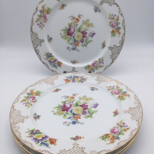 May include: Set of four white porcelain dinner plates with a floral design and gold trim. The plates have a delicate floral pattern with pink, yellow, and purple flowers. The gold trim is around the edge of the plates and features a delicate lace pattern.