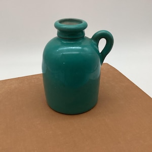 May include: A small, teal-colored ceramic jug with a handle. The jug has a rounded body and a narrow neck with a flared rim. The handle is a simple loop shape. The jug is sitting on a brown surface.