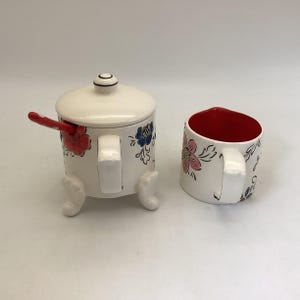 May include: A white ceramic sugar bowl with a lid and a matching mug. Both pieces are decorated with red, pink, and blue floral designs. The sugar bowl has a red spoon and is supported by three small feet.