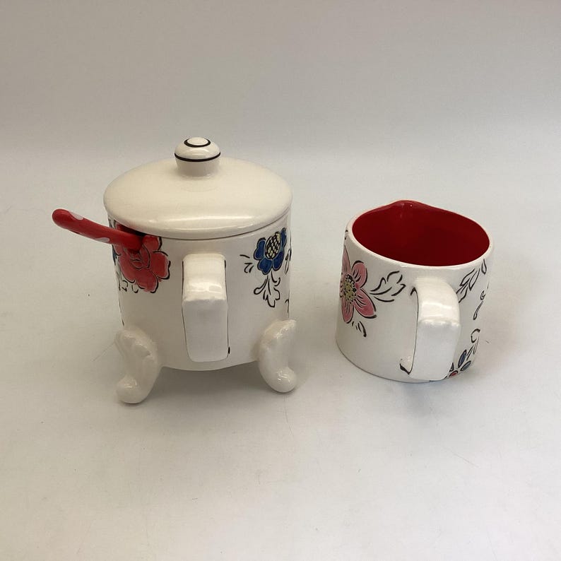 May include: A white ceramic sugar bowl with a lid and a matching mug. Both pieces are decorated with red, pink, and blue floral designs. The sugar bowl has a red spoon and is supported by three small feet.