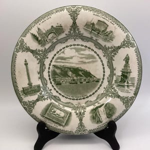 May include: A vintage white plate with a green border and illustrations of Quebec landmarks. The plate features images of the St. Louis Gate, Citadel, Wolfe Monument, and Montmorency Falls. The plate is displayed on a black stand.