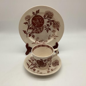 May include: A vintage cream-colored teacup and saucer set with a matching dinner plate. The set features a brown floral and bird design. The plate is propped up on a wooden stand. The teacup and saucer are in front of the plate.