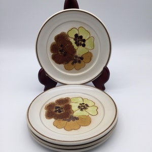 May include: A set of four off-white ceramic plates with a brown rim and a floral design. The flowers are brown, yellow, and tan. One plate is displayed on a dark brown plate stand.