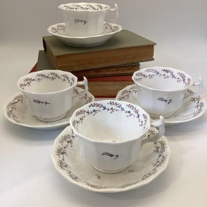 May include: A set of white teacups and saucers with a floral design. The cups have a unique handle shape and are paired with matching saucers. The floral pattern features purple, green, and blue accents. The teacups and saucers are arranged on a white surface.