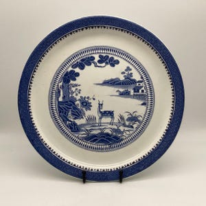 May include: A round, white ceramic plate with a blue and white Asian-inspired design. The plate features a central scene of a deer, trees, and a building, surrounded by a patterned border. The outer rim has a geometric design.