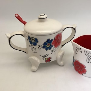 May include: A white ceramic sugar bowl with a lid, handles, and three feet. The bowl is decorated with red and blue floral designs and a red spoon. A matching creamer is visible to the right.