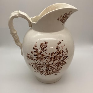May include: Cream-colored ceramic pitcher with a bulbous body and a decorative handle. The pitcher features a brown floral pattern across the front and near the spout. The spout is curved, and the handle has a detailed design.