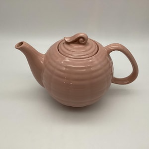 May include: A pink ceramic teapot with a round body, a curved spout, and a loop handle. The lid has a small, decorative handle. The teapot has a ridged texture around the body.