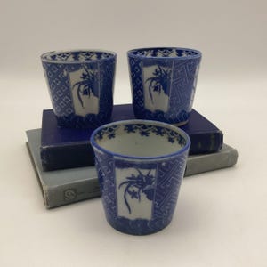 May include: Three blue and white ceramic cups with a floral design. The cups are cylindrical with a flared top and are decorated with a repeating geometric pattern and a central floral motif. The cups are stacked on top of each other.