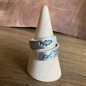 May include: Two silver-colored wrap rings displayed on a cone-shaped stand. One ring features a fish design, and the other has a mountain range. The rings are made of a shiny metal and are likely adjustable.