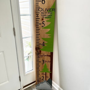 May include: A wooden growth chart with a green and brown forest theme. The chart is marked with numbers from 2 to 6. The text "OLIVER" is at the top of the chart, along with the date "MAY 17, 2022", weight "6 POUNDS, 5 OUNCES", and height "19 INCHES".