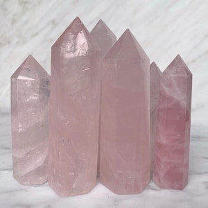 Pode incluir: Um grupo de seis pontas de cristal de quartzo rosa, também conhecidas como torres de quartzo rosa, dispostas em uma fileira sobre uma superfície de mármore branco.