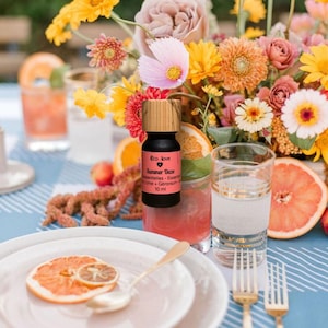 Puede incluir: Una botella negra de aceite esencial con tapa de madera, etiquetada "Eco. Love Summer Daze", está sobre una mesa con flores, bebidas y rodajas de fruta. La escena está ambientada sobre un mantel a cuadros azul y blanco.