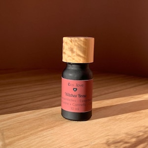 Peut inclure: Petite bouteille noire d'huile essentielle avec un bouchon en bois, étiquetée "Eco love" et "Witches Brew". La bouteille est posée sur une surface en bois avec des plantes séchées au premier plan. Le fond est un mur marron foncé.