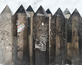 May include: Seven smoky quartz crystal points, each with a unique pattern of inclusions, are arranged in a row on a white surface. The points are all approximately the same size and shape, and they have a natural, unpolished finish.