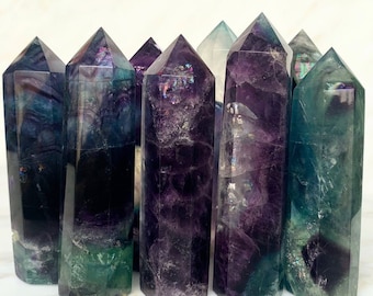 May include: A group of eight iridescent fluorite crystal points in shades of purple, green, and blue. The points are arranged in a row on a white surface.