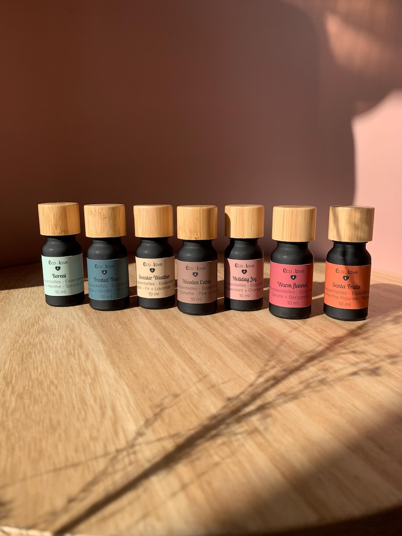 May include: Seven small glass bottles with wooden caps and labels. Each bottle contains essential oils with different scents and names: Boreal, Frosted Tree, Winter Weather, Wooden Cabin, Holiday Joy, Warm Flannel, and Santa Fruits. The bottles are arranged in a row on a wooden surface.