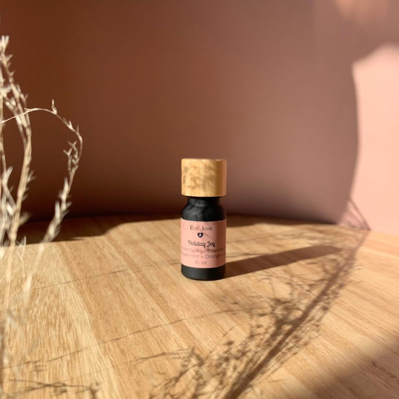 May include: A small black bottle with a wooden cap and a pink label that reads "Cool Love Holiday Joy" with a small heart symbol. The label also says "Essential Oil Blend" and "Cinnamon + Orange" and "10 ml".