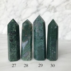 May include: Four green moss agate crystal points, numbered 27 to 30, arranged in a row on a white marble surface.