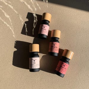 Peut inclure: Quatre petits flacons d'huiles essentielles noirs avec des bouchons en bois sont disposés sur une surface claire. Chaque flacon porte une étiquette avec la marque "Eco. love" et un nom de parfum différent, comme "Romance" et "Silk". Les flacons font 10 ml chacun.