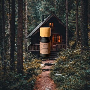 May include: A small, dark glass bottle with a wooden cap, labeled "Wooden Cabin" essential oil, sits in front of a rustic cabin in a forest. The bottle is 10 ml. The background features a path leading to the cabin, surrounded by tall trees.