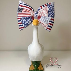 May include: A decorative goose figurine with a large patriotic bow. The bow features a patchwork design with red, white, and blue fabrics, including an American flag pattern. The goose has a white body, yellow beak, and green and yellow feet.