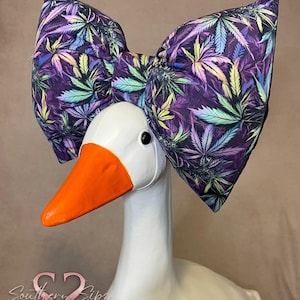 May include: A white goose statue with an orange beak and a large bow. The bow features a colorful cannabis leaf pattern on a purple background. The goose has a white body and a black eye.