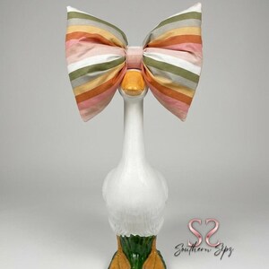 May include: A white ceramic goose figurine with a large striped bow in shades of pink, orange, green, and white. The goose has orange feet and a green base. The bow is centered on the goose's neck.