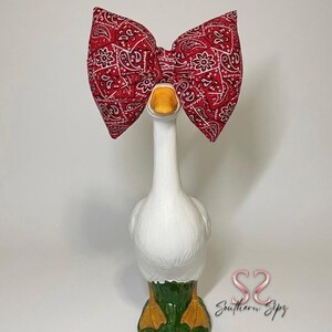 May include: A white goose figurine with a red paisley bow. The goose has a yellow beak and feet, with green and yellow painted details. The bow is a large, decorative accent.