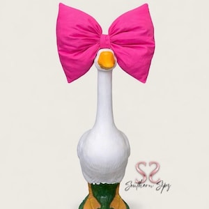 May include: A whimsical white goose statue with a bright pink bow. The goose has a yellow beak and feet, and is standing on a green base. The statue is a decorative item.
