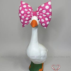 May include: A white goose figurine with an orange beak and feet, wearing a large pink bow with white polka dots. The goose stands on a green base. The figurine has a whimsical and decorative design.