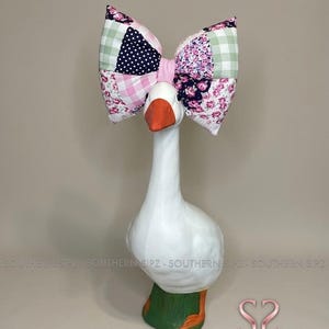 May include: A white goose figurine with an orange beak and green feet, adorned with a large patchwork bow. The bow features pink, white, navy, and floral patterns. The figurine has the text "SOUTHERN SIPZ" on the bottom.