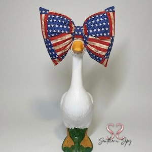 May include: A white goose figurine with a large bow tie featuring a patriotic design of red stripes and blue stars. The goose has a yellow beak and feet, with green grass at the base. The bow tie is approximately 10 inches wide.