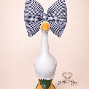 May include: A white goose figurine with a large navy and white gingham bow. The goose has an orange beak and feet, with green and orange accents at the base. The figurine is on a light pink background.