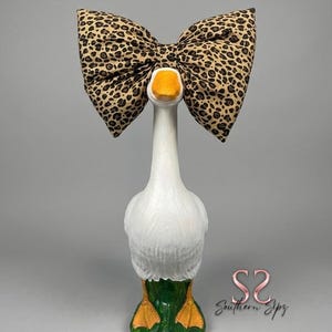 May include: A white goose statue with an orange beak and feet, wearing a large leopard print bow. The base of the goose is green. The statue has a textured surface and is set against a gray background.