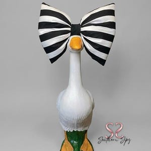 May include: A white goose statue with a large black and white striped bow. The goose has an orange beak and feet, with green and orange accents. The statue has the text "Southern Sipz" in the bottom right corner.
