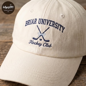 May include: A cream-colored baseball cap featuring dark blue embroidered text that reads Briar University Hockey Club. The design includes two crossed dark blue hockey sticks tied with a light blue bow and a small dark blue heart below them.