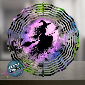 May include: A decorative metal wind spinner featuring a silhouette of a witch on a broomstick against a purple and blue watercolor background. The design includes spiders and a full moon, with a wavy, layered metal frame. The text "PLANET CRAFTY" is visible.