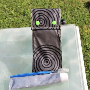 May include: A black and grey toiletry bag with a circular pattern. The bag has two green buttons and a small black tag with the text "7711". A blue toothbrush and a tube of toothpaste are on a white surface in front of the bag.