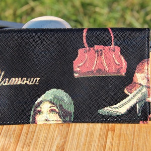 Peut inclure: Portefeuille en tissu noir avec une inscription dorée qui dit "Glamour". Le portefeuille présente un sac à main rouge, une chaussure de femme et un visage de femme.