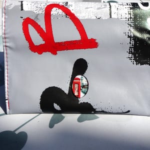 Op de afbeelding: Een grijze clutch met een rood abstract ontwerp en een zwarte inktvlek. Een klein rond venster toont een rode telefooncel. De randen zijn afgewerkt met rode stiksels.