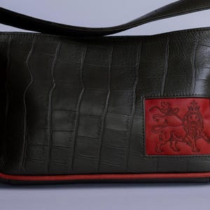 May include: A dark green leather handbag with a red trim and a red leather patch featuring a lion design.