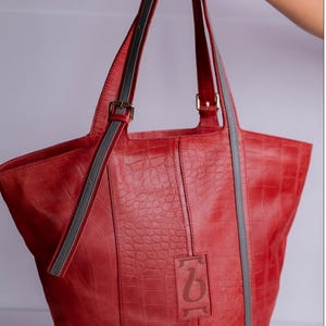 Puede incluir: Un bolso tote de cuero rojo con correas grises y un patrón de cocodrilo en relieve. El bolso tiene un logotipo en la parte delantera que dice "b".