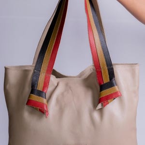 Peut inclure: Un sac cabas en cuir beige avec des lanières en cuir rouge, noir et marron. Les lanières sont nouées en un nœud au sommet du sac. Le sac a un patch rouge avec un motif noir sur le devant.