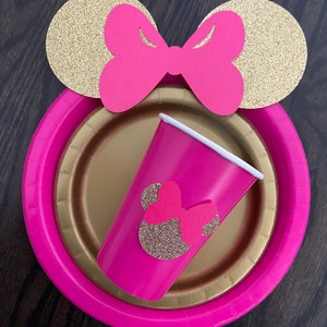 Puede incluir: Suministros para fiestas con tem&aacute;tica de Minnie Mouse en rosa y dorado. Un plato de papel rosa con borde dorado, una taza rosa con una cabeza de Minnie Mouse de purpurina dorada y orejas de Minnie Mouse de purpurina dorada con un lazo rosa.