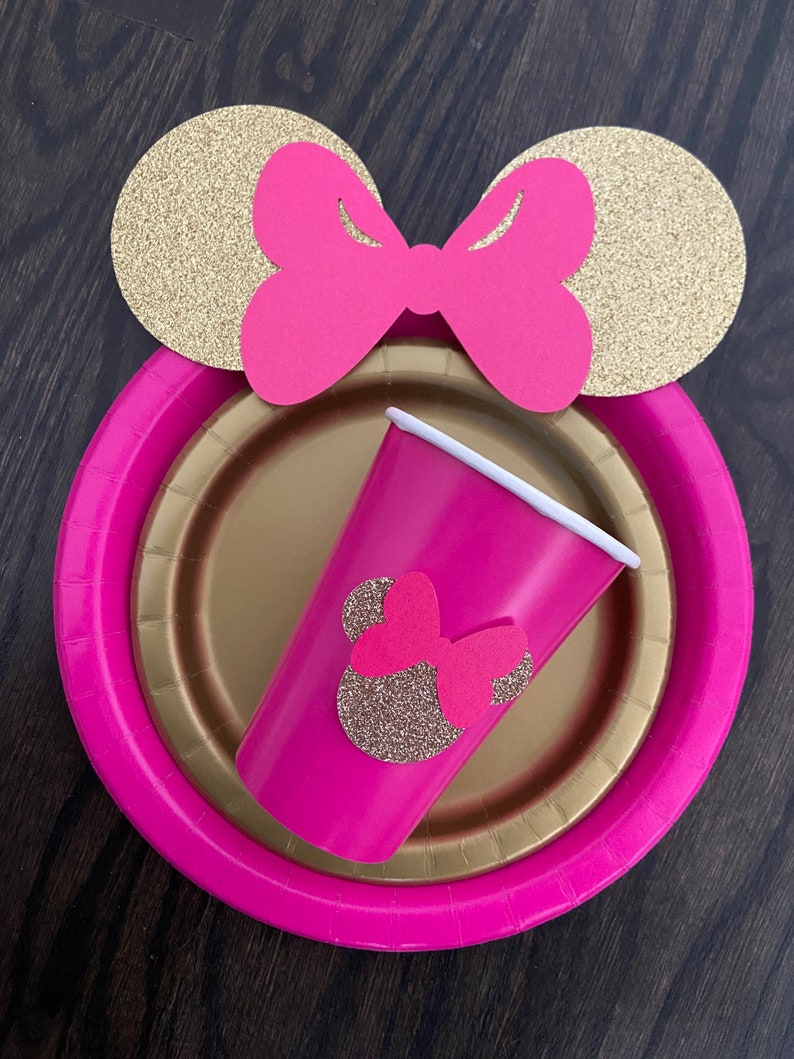 Puede incluir: Suministros para fiestas con tem&aacute;tica de Minnie Mouse en rosa y dorado. Un plato de papel rosa con borde dorado, una taza rosa con una cabeza de Minnie Mouse de purpurina dorada y orejas de Minnie Mouse de purpurina dorada con un lazo rosa.