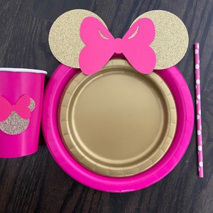 Puede incluir: Art&iacute;culos de fiesta de tem&aacute;tica Minnie Mouse en rosa y dorado, que incluyen una taza, un plato y una pajita. La taza y el plato tienen una silueta de Minnie Mouse con un lazo rosa. La pajita tiene lunares blancos.