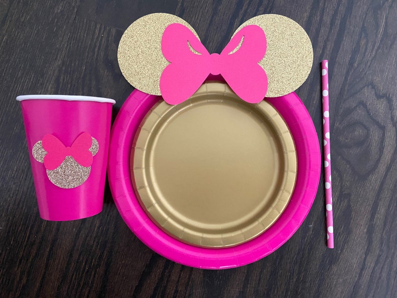 Puede incluir: Art&iacute;culos de fiesta de tem&aacute;tica Minnie Mouse en rosa y dorado, que incluyen una taza, un plato y una pajita. La taza y el plato tienen una silueta de Minnie Mouse con un lazo rosa. La pajita tiene lunares blancos.